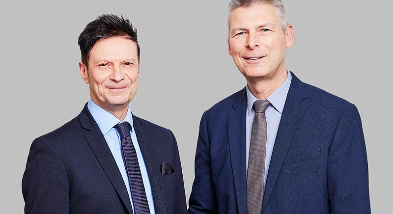 Portrait von Jörg Kamphaus und Dr. Thomas Becker in Anzügen vor einem neutralen grauen Hintergrund.