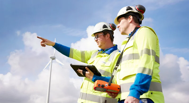 Zwei Thyssengas-Mitarbeiter mit Messtechnik vor einer Windkraftanlage, Symbol für den zukünftigen Energietransport.