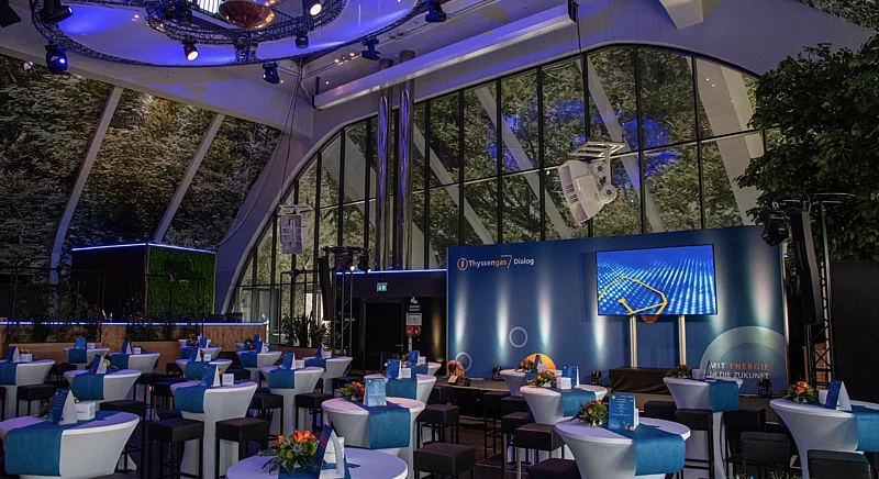 Modern eingerichteter Veranstaltungsraum für den Thyssengas Dialog mit Bühne und blau-weißen Stehtischen.