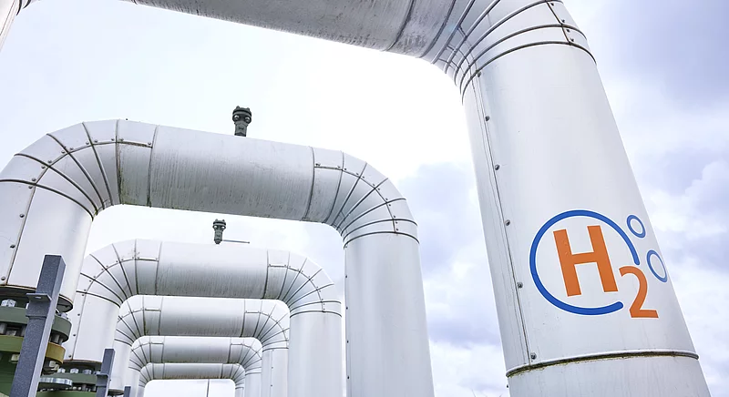 Große Pipeline mit H2-Logo für den Wasserstofftransport, im Hintergrund Windräder als Symbol für die Energiewende.