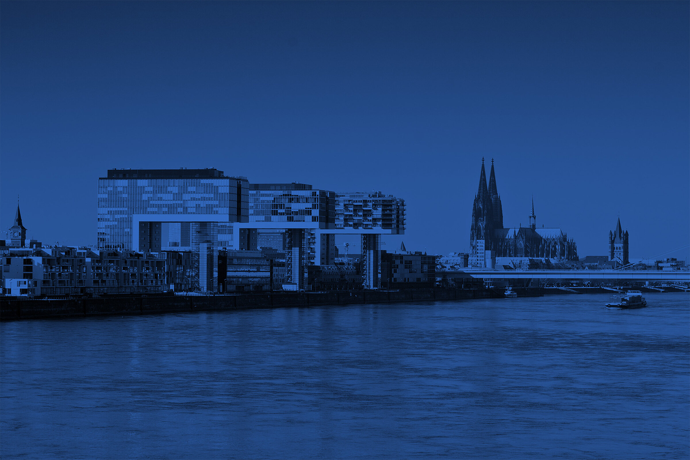 Blau getöntes Stimmungsbild der Kölner Skyline mit Kranhäusern und Dom am Rhein.