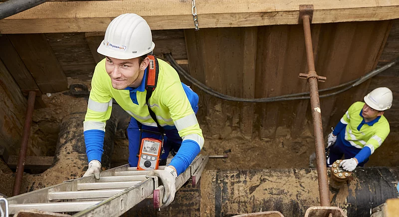 Zwei Thyssengas-Monteure bei Leitungsbauarbeiten in einer Baugrube; einer steigt auf einer Leiter nach oben.