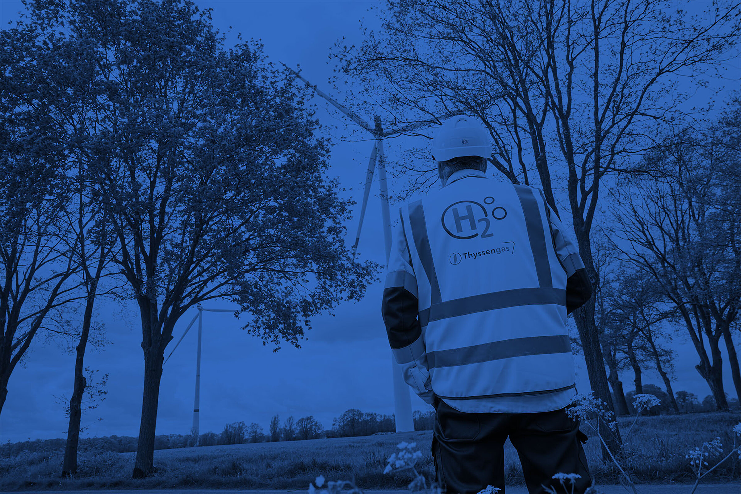 Thyssengas-Mitarbeiter mit H2-Logo auf der Weste blickt auf Windräder, Symbol für Jobs in der Energiewende.