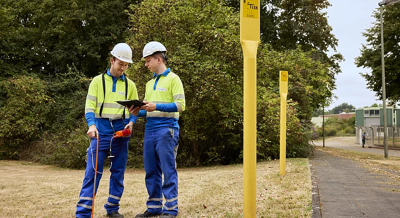 Zwei Thyssengas-Techniker bei der Inspektion einer Gasleitung mit Messgerät und Tablet an gelben Markierungspfählen.