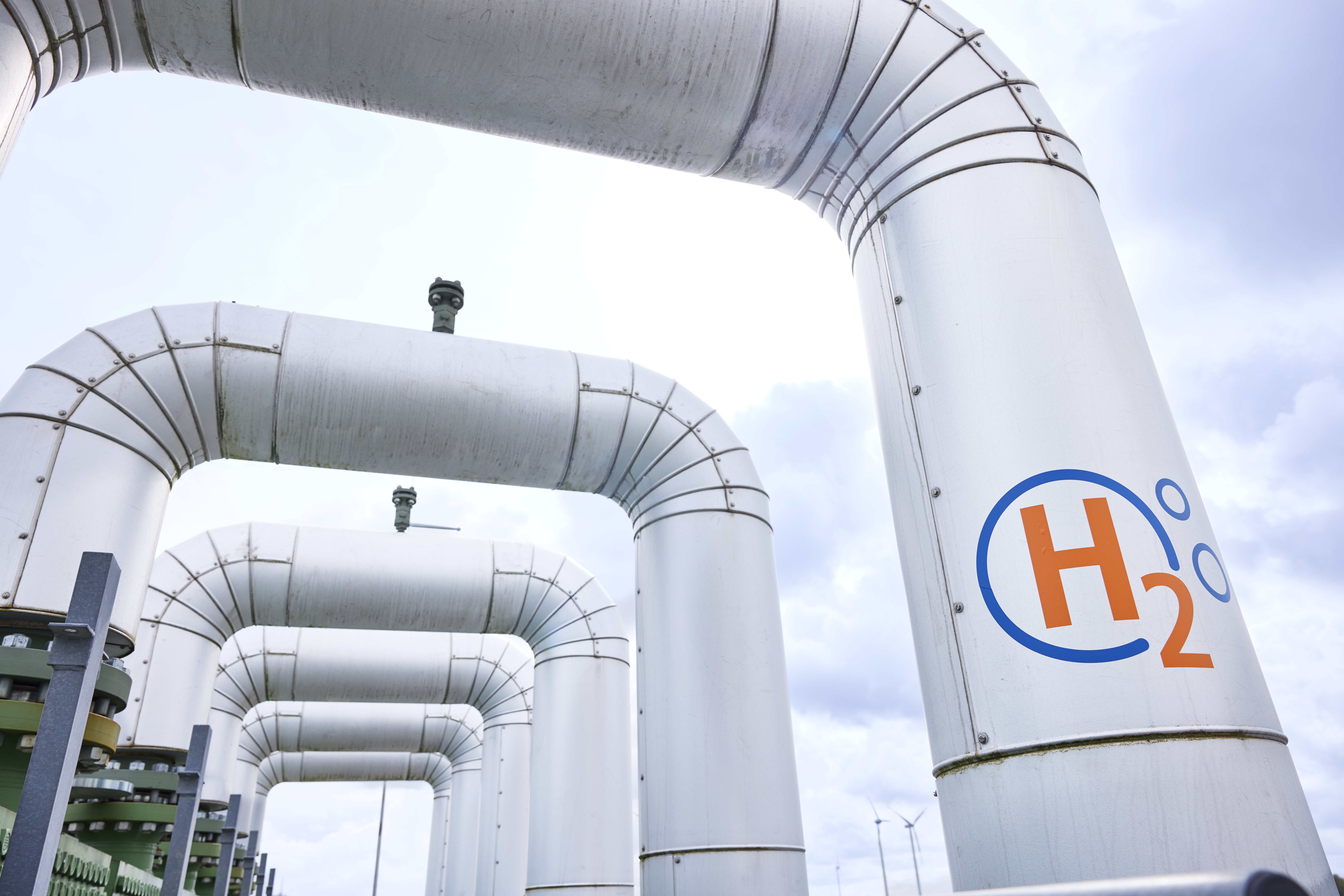 Große Pipeline mit H2-Logo für den Wasserstofftransport, im Hintergrund Windräder als Symbol für die Energiewende.