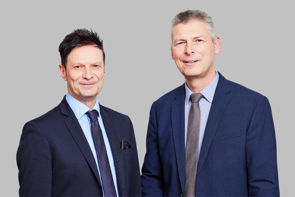 Portrait von Jörg Kamphaus und Dr. Thomas Becker in Anzügen vor einem neutralen grauen Hintergrund.