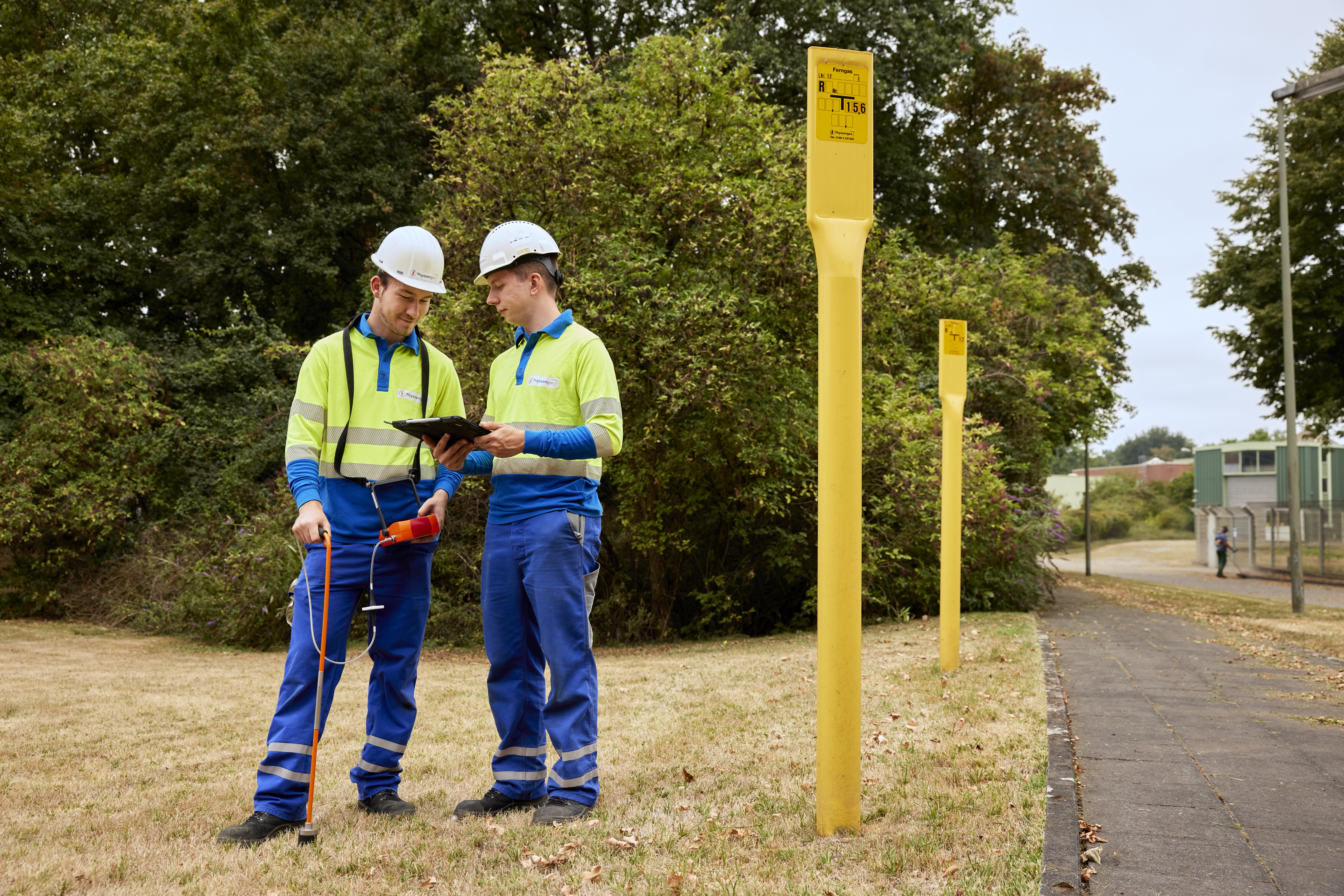 Zwei Thyssengas-Techniker bei der Inspektion einer Gasleitung mit Messgerät und Tablet an gelben Markierungspfählen.